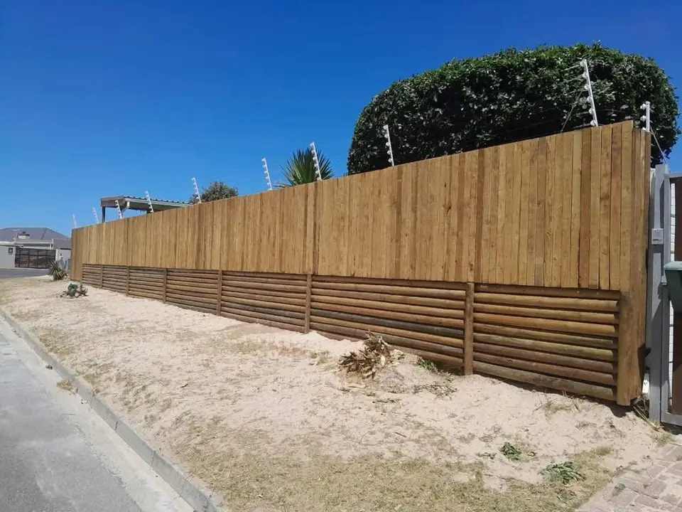Tall vertical timber fence on a horizontal pole base along a sandy verge